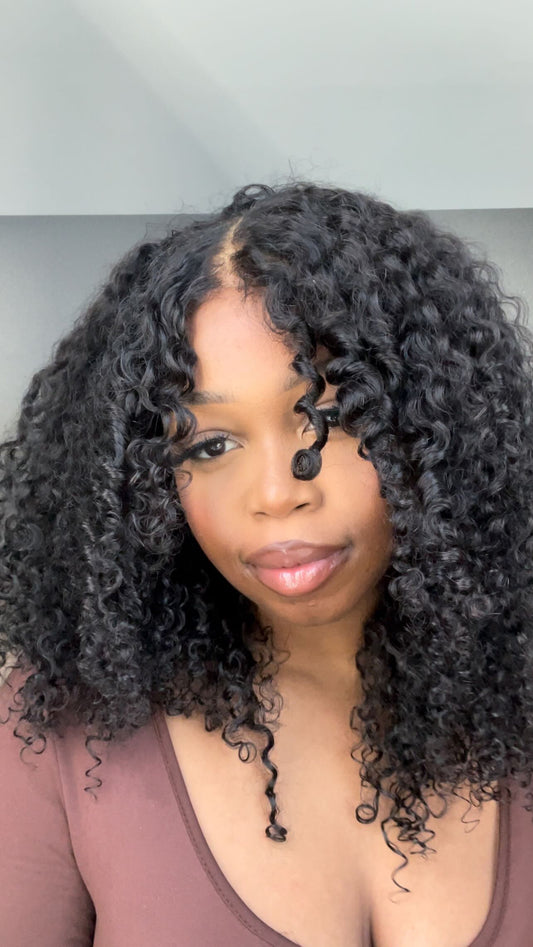 A close-up portrait of a woman with voluminous, glossy, black curly hair (likely a wig or hair extension) with a deep side part, wearing a mauve-brown scoop neck top.